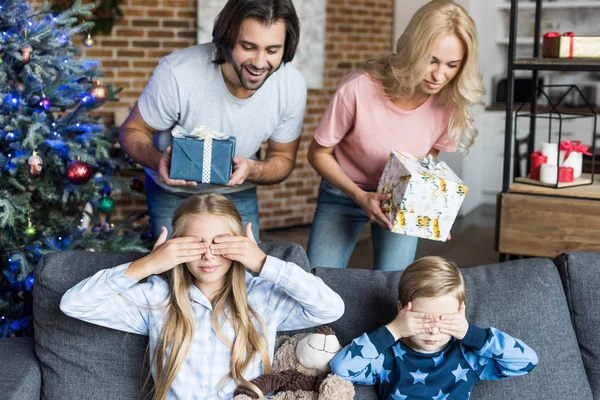 Родители преподносят рождественские подарки очаровательным детям в пижамах закрывая глаза — стоковое фото