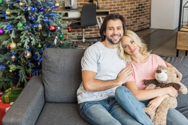 Vue grand angle de beau jeune couple heureux assis sur le canapé et souriant à la caméra à Noël — Photo de stock