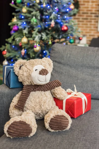 Nahaufnahme von Teddybär mit Geschenkschachteln auf Sofa und geschmücktem Weihnachtsbaum dahinter — Stockfoto