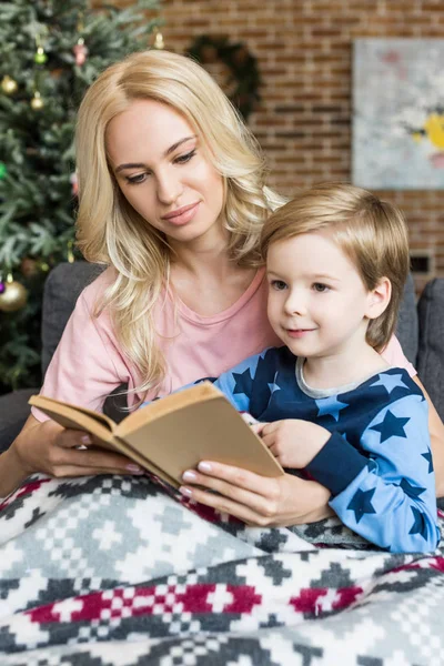 Beautiful young mother and adorable smiling son reading book together at christmas time — Stock Photo
