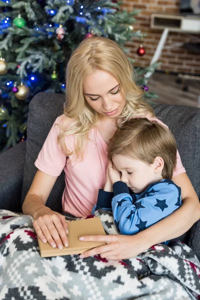 Jovem mãe segurando livro e olhando para bonito pequeno filho em pijama dormindo no sofá no Natal tempo — Fotografia de Stock