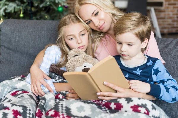 Счастливая молодая мама читает книжку с симпатичными детьми в пижамах в разгар праздника — стоковое фото
