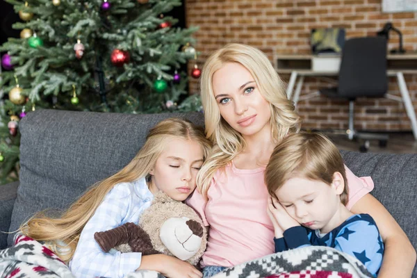 Молодая мама смотрит в камеру, сидя с милыми маленькими цыплятами в пижамах, спящими на диване в рождественское время — стоковое фото