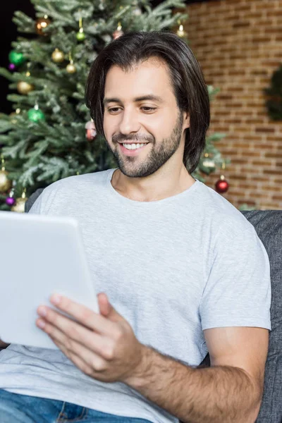 Joven sonriente usando tableta digital mientras está sentado en el sofá en Navidad - foto de stock