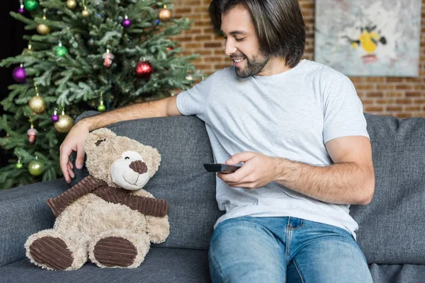 Smiling man holding remote controller and looking at teddy bear on couch at christmas time — Stock Photo