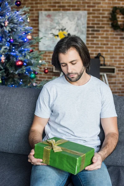 Pensive young man sitting on couch and looking at christmas present — Stock Photo