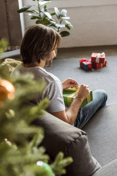 Enfoque selectivo del hombre sentado en el suelo cerca del árbol de Navidad y la apertura de la caja de regalo - foto de stock