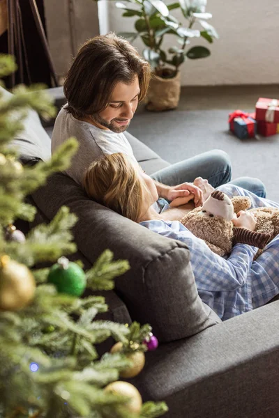 Vista de ángulo alto de padre feliz mirando a linda hija en pijama sosteniendo oso de peluche y durmiendo en el sofá en tiempo de Navidad - foto de stock