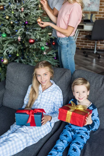 Vista de ángulo alto de niños felices en pijama sosteniendo regalos de Navidad y sonriendo a la cámara mientras los padres decoran el árbol de Navidad detrás - foto de stock