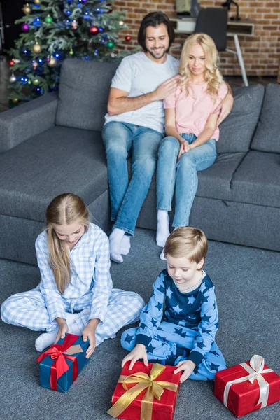 Vista de ángulo alto de los niños lindos en pijamas abriendo regalos de Navidad mientras los padres felices se sientan detrás - foto de stock