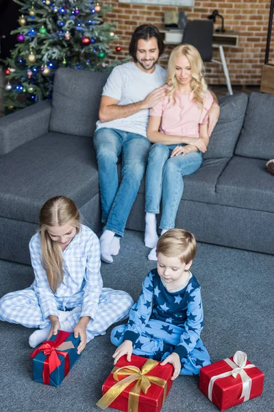 Високий кут зору чарівних дітей у піжамах, що відкривають різдвяні подарунки, а щасливі батьки сидять позаду — стокове фото