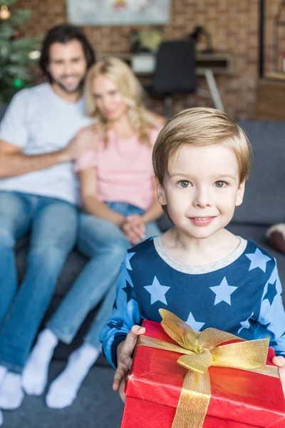 Adorabile bambino felice in pigiama che tiene il regalo di Natale e sorride alla macchina fotografica mentre i genitori seduti dietro — Foto stock