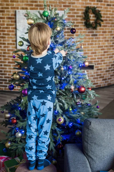 Back view of cute little boy in pajamas decorating christmas tree — Stock Photo