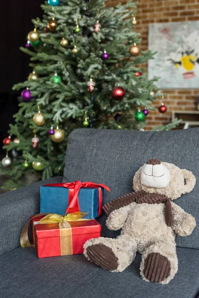 Oso de peluche con cajas de regalo en el sofá y árbol de Navidad decorado detrás - foto de stock