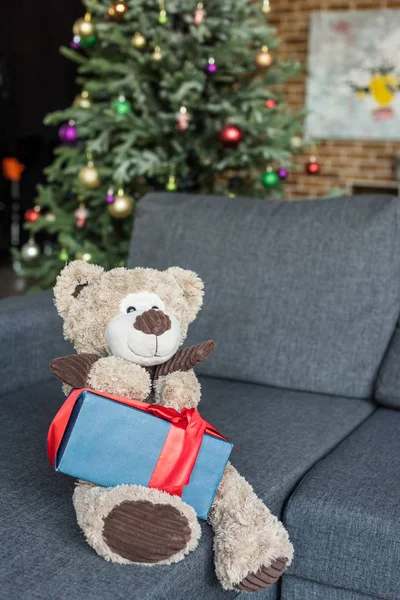 Плюшевий ведмідь з подарунком на дивані і прикрашена ялинка позаду — стокове фото