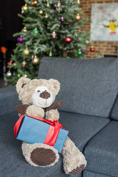 Teddy bear with present on couch and decorated christmas tree behind — Stock Photo