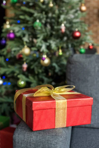 Nahaufnahme von roter Geschenkbox mit goldener Schleife auf Couch und geschmücktem Weihnachtsbaum dahinter — Stockfoto