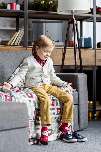 Милый улыбающийся ребенок сидит на диване и смотрит на обувь — стоковое фото