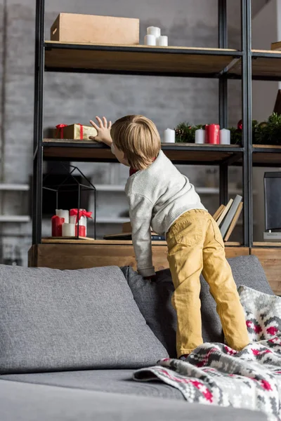 Vista posterior del niño de pie en el sofá y alcanzar el regalo de Navidad - foto de stock