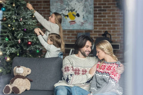 Selective focus of happy parents sitting in sofa while children decorating christmas tree behind — Stock Photo