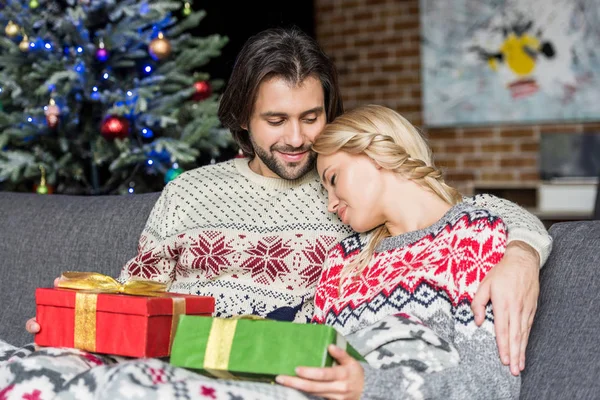 Hermosa pareja joven sosteniendo regalos de Navidad y sentados juntos en el sofá - foto de stock