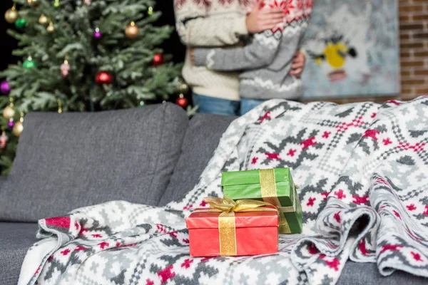 Рождественские подарки на диване и молодая пара, стоящая возле рождественской елки позади — стоковое фото
