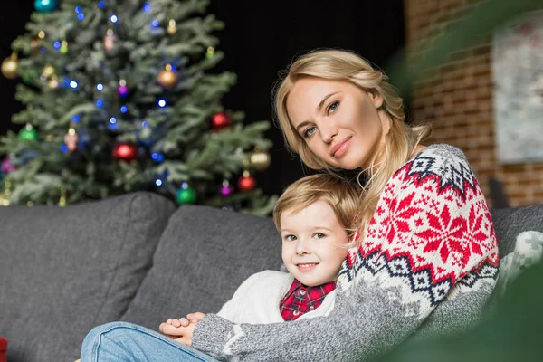 Mère heureuse et mignon petit fils assis ensemble sur le canapé et souriant à la caméra à Noël — Photo de stock
