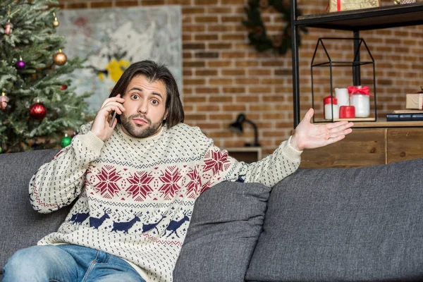 Мужчина в свитере разговаривает по смартфону и смотрит в камеру, сидя на диване в канун Рождества — стоковое фото