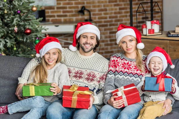 Heureux famille dans santa chapeaux tenant cadeaux de Noël et souriant à la caméra — Photo de stock