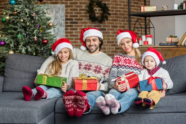 Heureux famille dans santa chapeaux tenant cadeaux de Noël et assis ensemble à la maison — Photo de stock