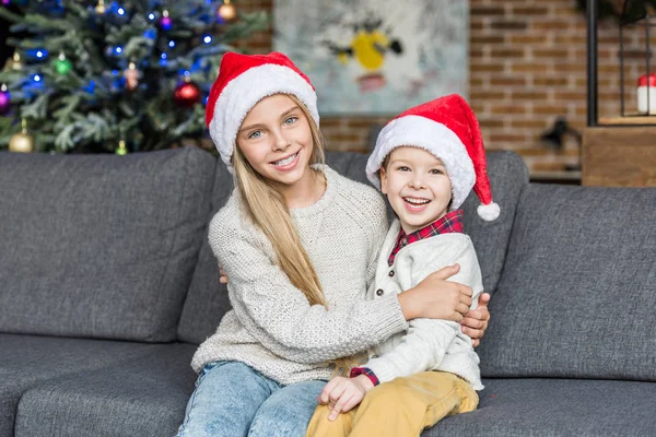 Lindo feliz niños en santa sombreros sentado juntos en sofá y sonriendo a la cámara - foto de stock