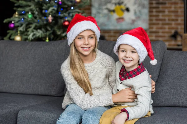 Adorables enfants heureux dans chapeaux de Père Noël assis ensemble sur le canapé et souriant à la caméra — Photo de stock