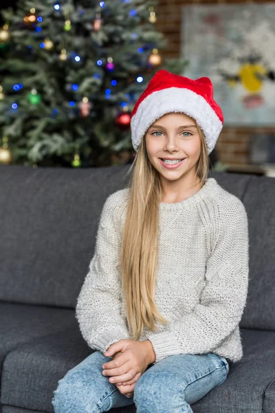 Beautiful kid in santa hat sitting on couch and smiling at camera — Stock Photo