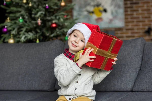 Entzückendes lächelndes Kind mit Weihnachtsmütze und Weihnachtsgeschenk zu Hause — Stockfoto