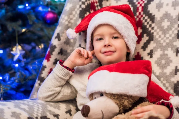 Cute little boy in santa hat holding teddy bear and smiling at camera at christmas time — Stock Photo