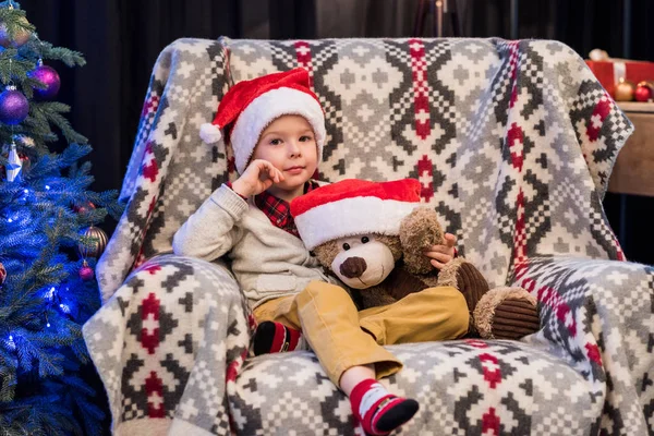 Очаровательный маленький мальчик держит плюшевого мишку в шляпе Санты и улыбается в камеру на Рождество — стоковое фото