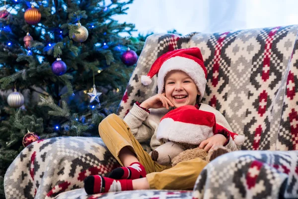 Adorable cheerful little boy holding teddy bear in santa hat and smiling at camera — Stock Photo