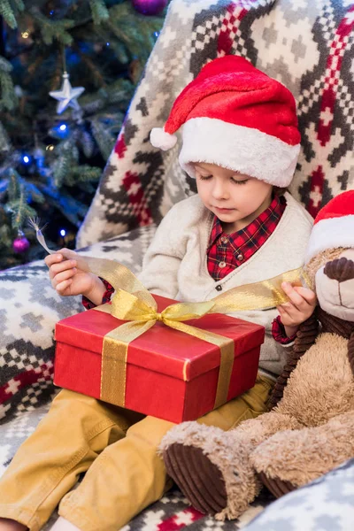 Adorable little boy in santa hat opening christmas present — Stock Photo