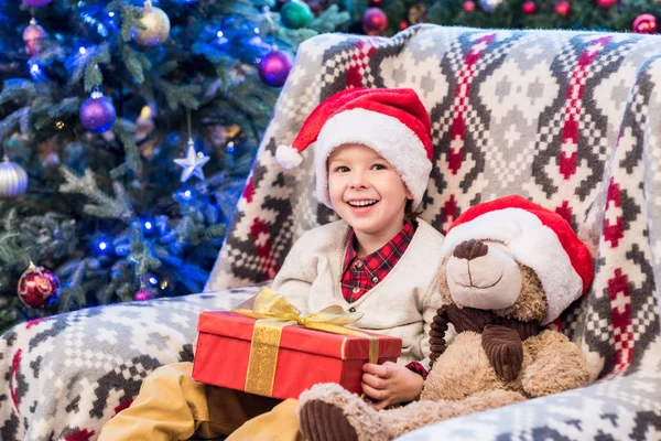 Happy little boy in santa hat holding christmas present and smiling at camera — Stock Photo
