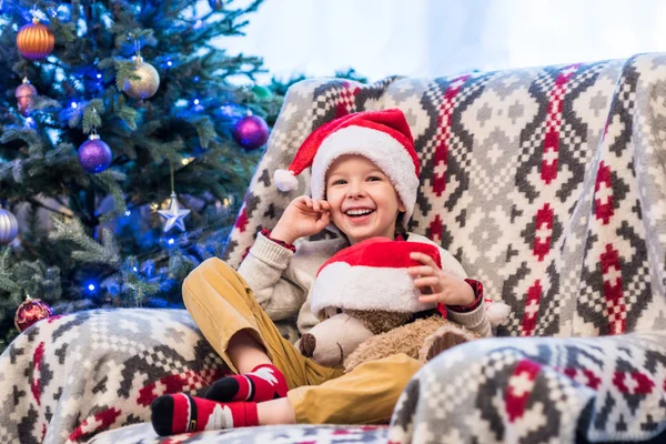 Happy little boy holding teddy bear in santa hat and smiling at camera — Stock Photo