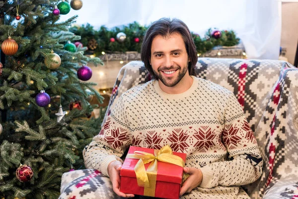 Glücklicher junger Mann hält Weihnachtsgeschenk in der Hand und lächelt in die Kamera — Stockfoto