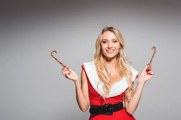 Heureuse jeune femme en robe de Noël montrant des bâtons de bonbons rayés isolé sur fond gris — Photo de stock