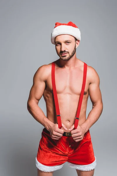 Уверенно сексуальный мужчина в рождественской шляпе и шортах держа подтяжки изолированы на сером фоне — стоковое фото