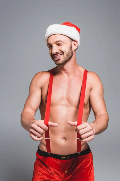 Lächelnder muskulöser Mann mit Weihnachtsmütze zwinkert und hält Hosenträger isoliert auf grauem Hintergrund — Stockfoto