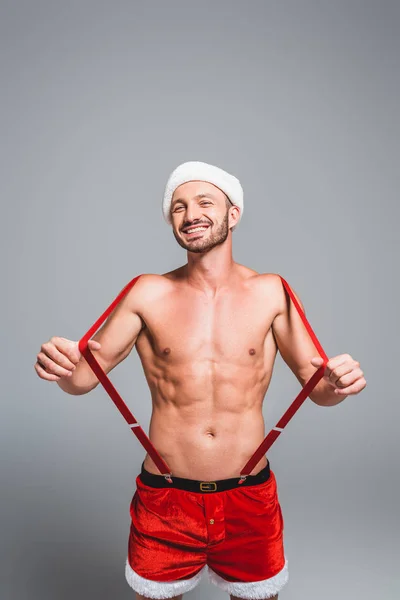 Lachender, muskulöser Mann mit Weihnachtsmütze und kurzen Hosen, der Hosenträger isoliert auf grauem Hintergrund hält — Stockfoto