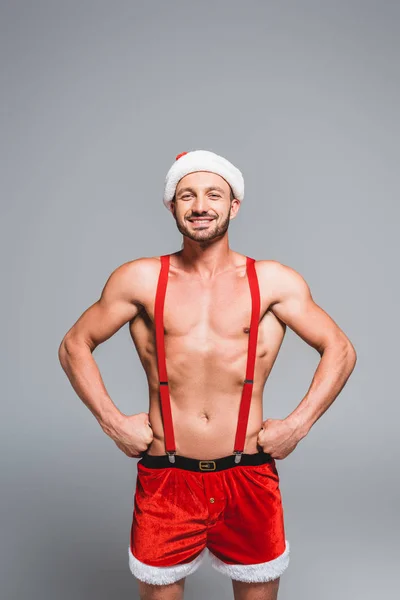 Glücklicher muskulöser, hemdloser Mann mit Weihnachtsmütze und Shorts, stehend mit den Händen auf der Taille isoliert auf grauem Hintergrund — Stockfoto