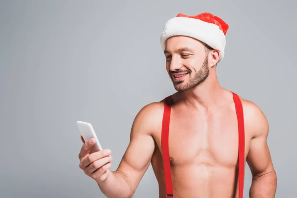 Hombre musculoso en sombrero de Navidad mirando teléfono inteligente aislado sobre fondo gris - foto de stock
