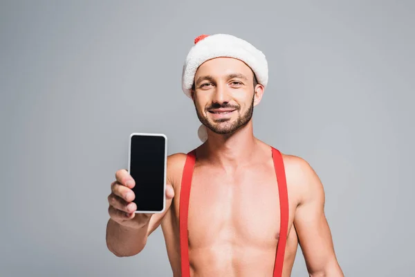 Щасливий м'язистий чоловік у різдвяному капелюсі, що показує смартфон з порожнім екраном ізольовано на сірому фоні — стокове фото