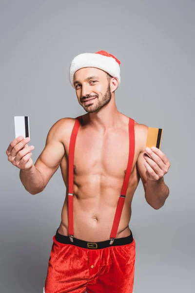 Schöner muskulöser Mann mit Weihnachtsmütze, der Kreditkarten isoliert auf grauem Hintergrund zeigt — Stockfoto