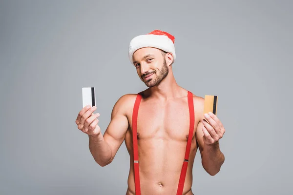 Young sexy muscular man in christmas hat showing credit cards isolated on grey background — Stock Photo